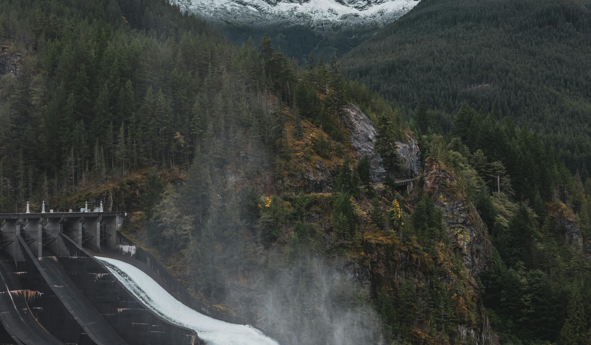 Concrete dam wall with overflowing spillway, snow-capped mountains and pine forest behind